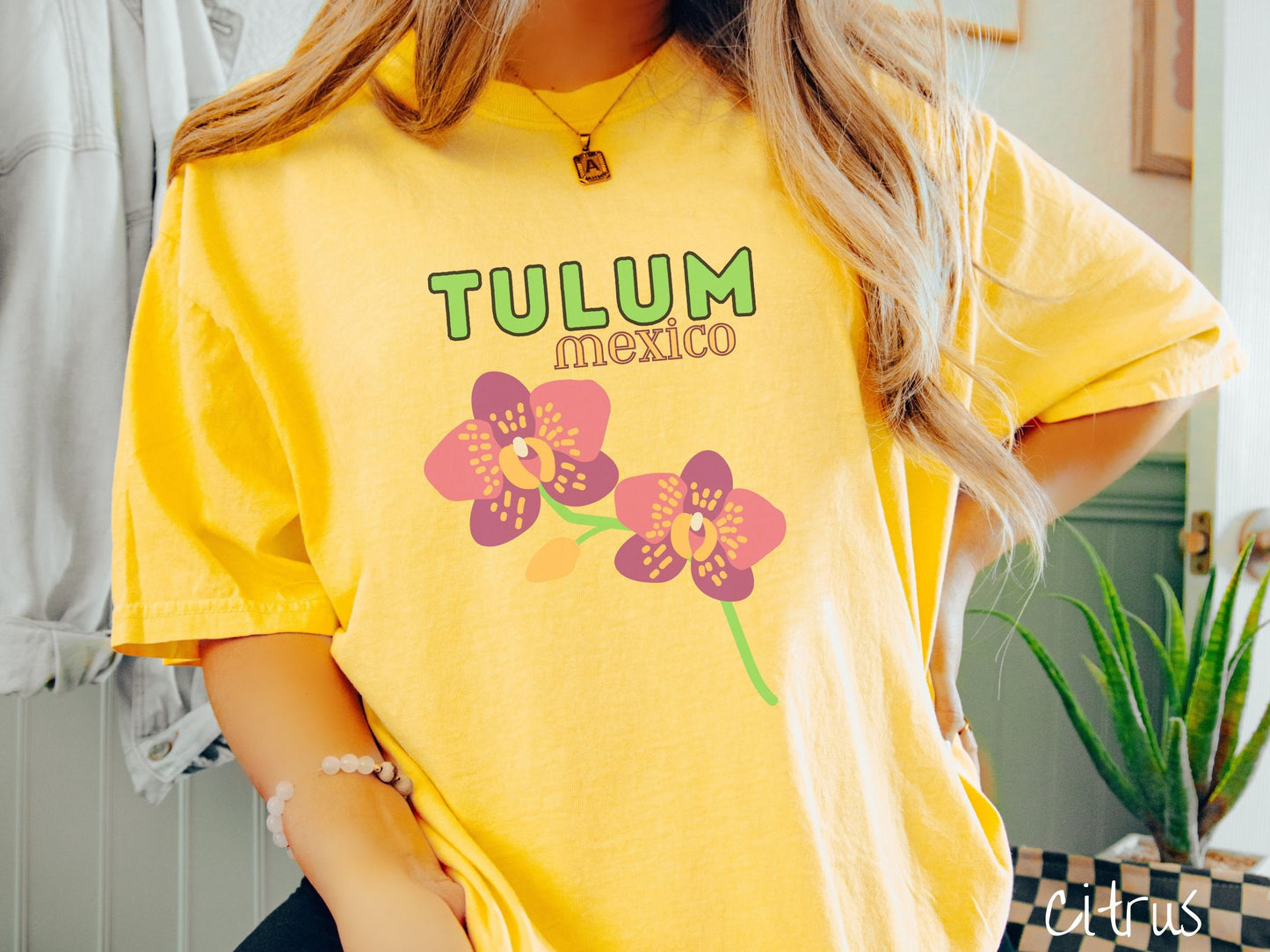 A woman wearing a vintage, citrus colored shirt with the text Tulum Mexico in green and yellow font, respectively, at the top and below it are two beautiful pink and purple flowers with green stems.