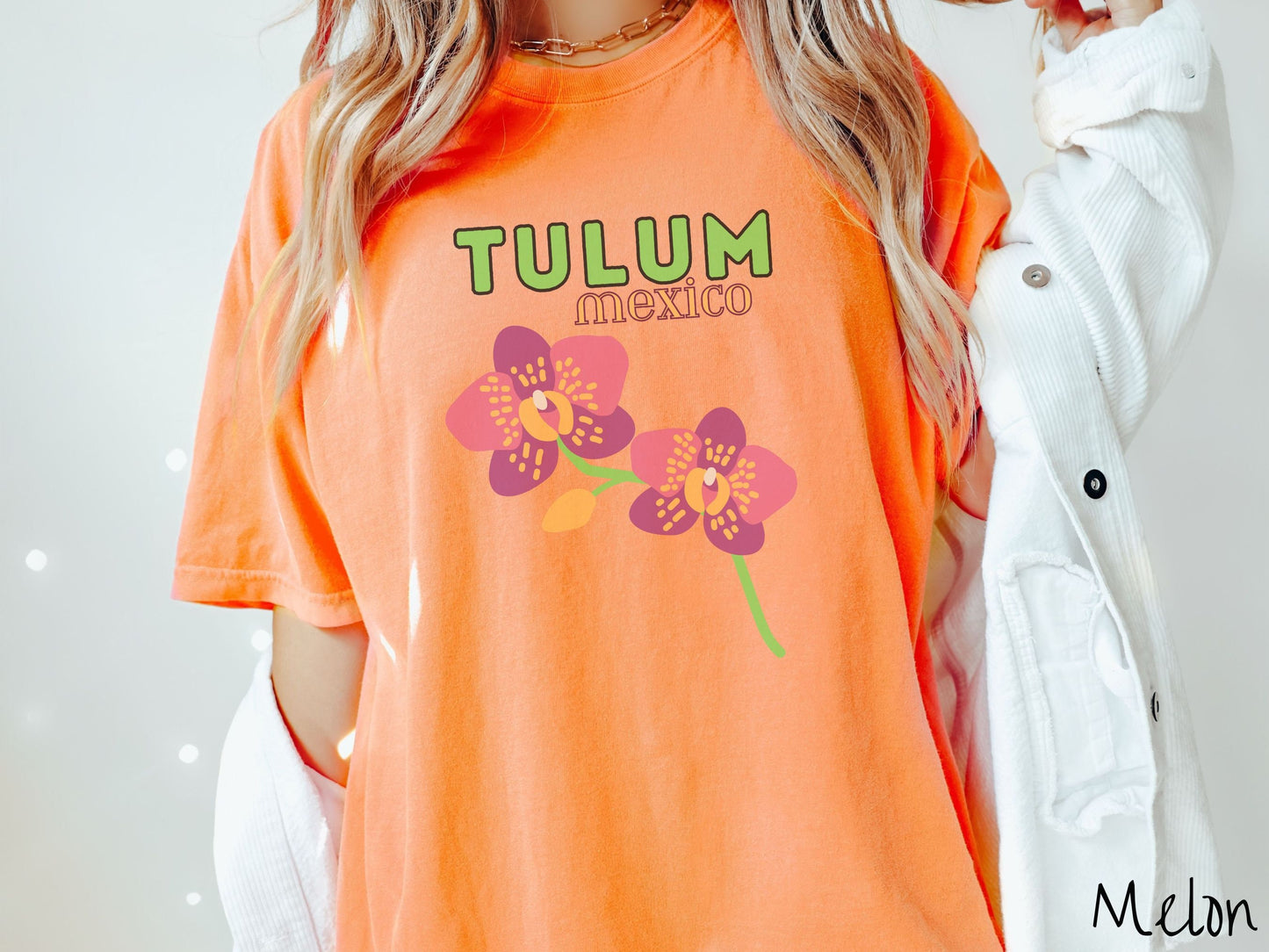 A woman wearing a vintage, melon colored shirt with the text Tulum Mexico in green and yellow font, respectively, at the top and below it are two beautiful pink and purple flowers with green stems.