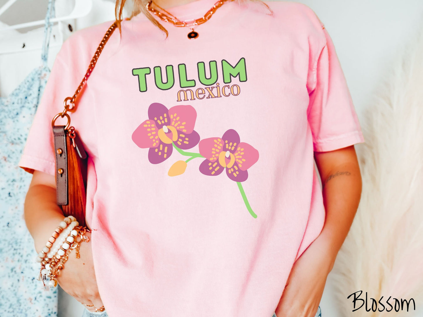 A woman wearing a vintage, blossom colored shirt with the text Tulum Mexico in green and yellow font, respectively, at the top and below it are two beautiful pink and purple flowers with green stems.