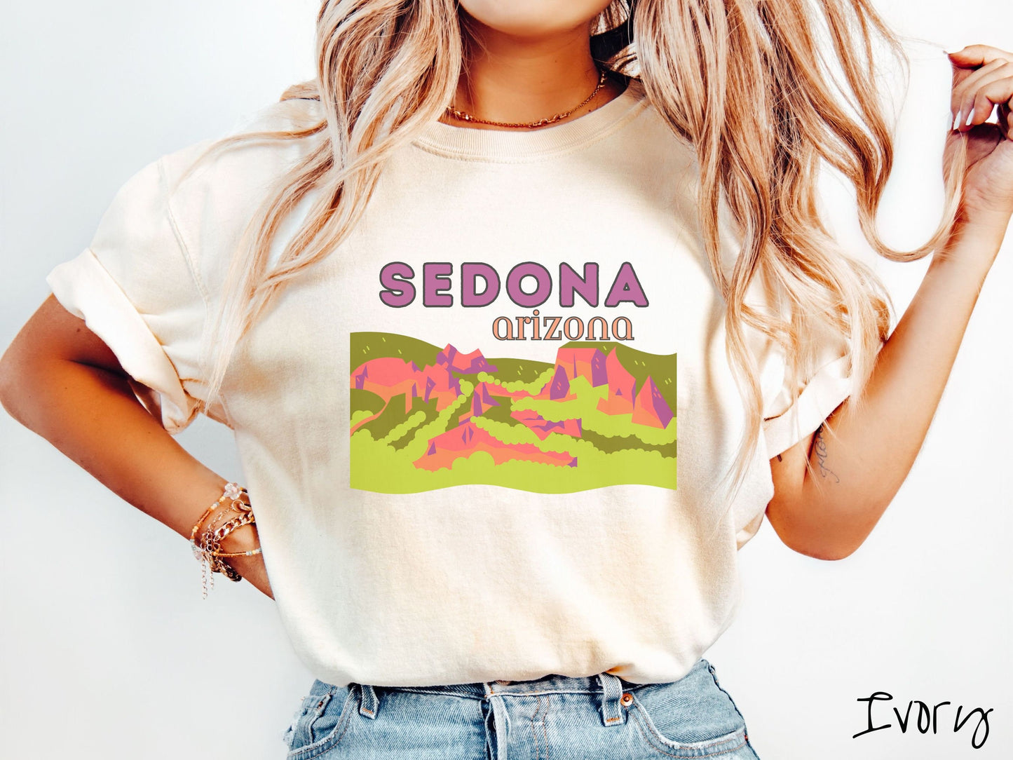 A woman wearing a vintage, ivory colored shirt with the text Sedona Arizona in purple and orange font, respectively, and below that is a picture of red rock canyons and green meadows.