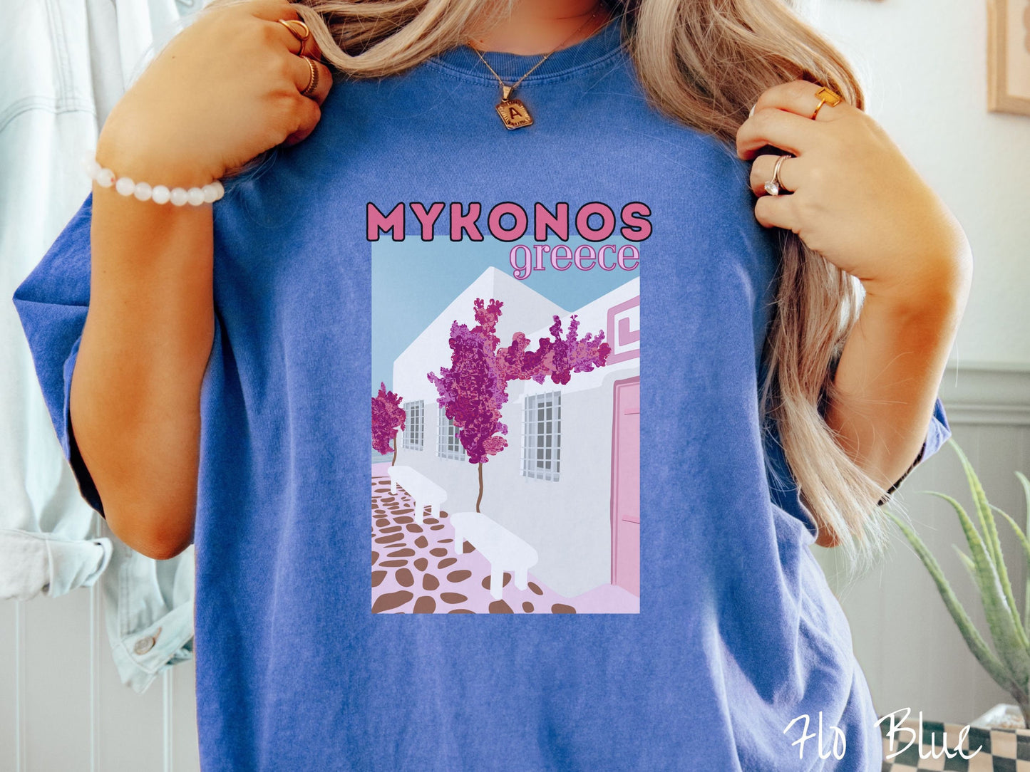 A woman wearing a vintage, flo blue colored shirt with the text Mykonos Greece at the top, and beneath is a picture of Cycladic architecture, a beautiful white home with purple trees in the front yard, and a pink driveway with brown stone.