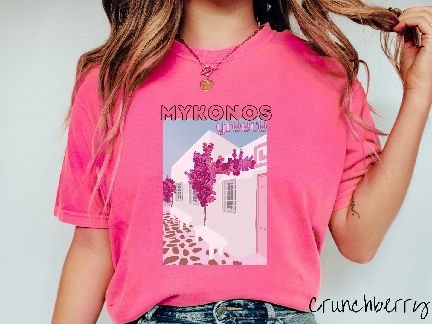 A woman wearing a vintage, crunchberry colored shirt with the text Mykonos Greece at the top, and beneath is a picture of Cycladic architecture, a beautiful white home with purple trees in the front yard, and a pink driveway with brown stone.