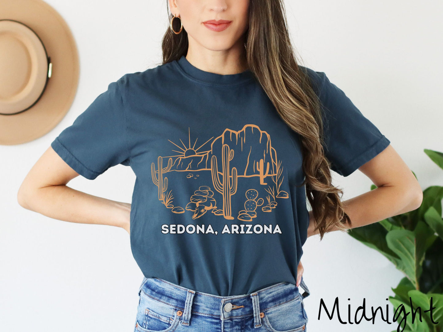 A woman wearing a cute, vintage midnight colored Comfort Colors t-shirt with text Sedona, Arizona in white font. Above is a black outline of a desert landscape with the sun and mountains in the background, and cacti and rocks in the foreground.