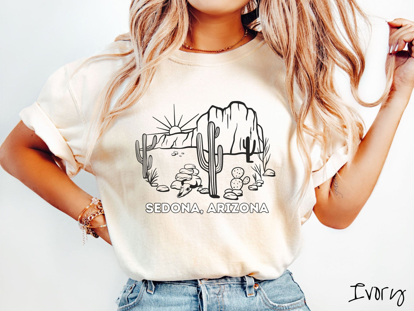 A woman wearing a cute, vintage ivory colored Comfort Colors t-shirt with text Sedona, Arizona in white font. Above is a black outline of a desert landscape with the sun and mountains in the background, and cacti and rocks in the foreground.