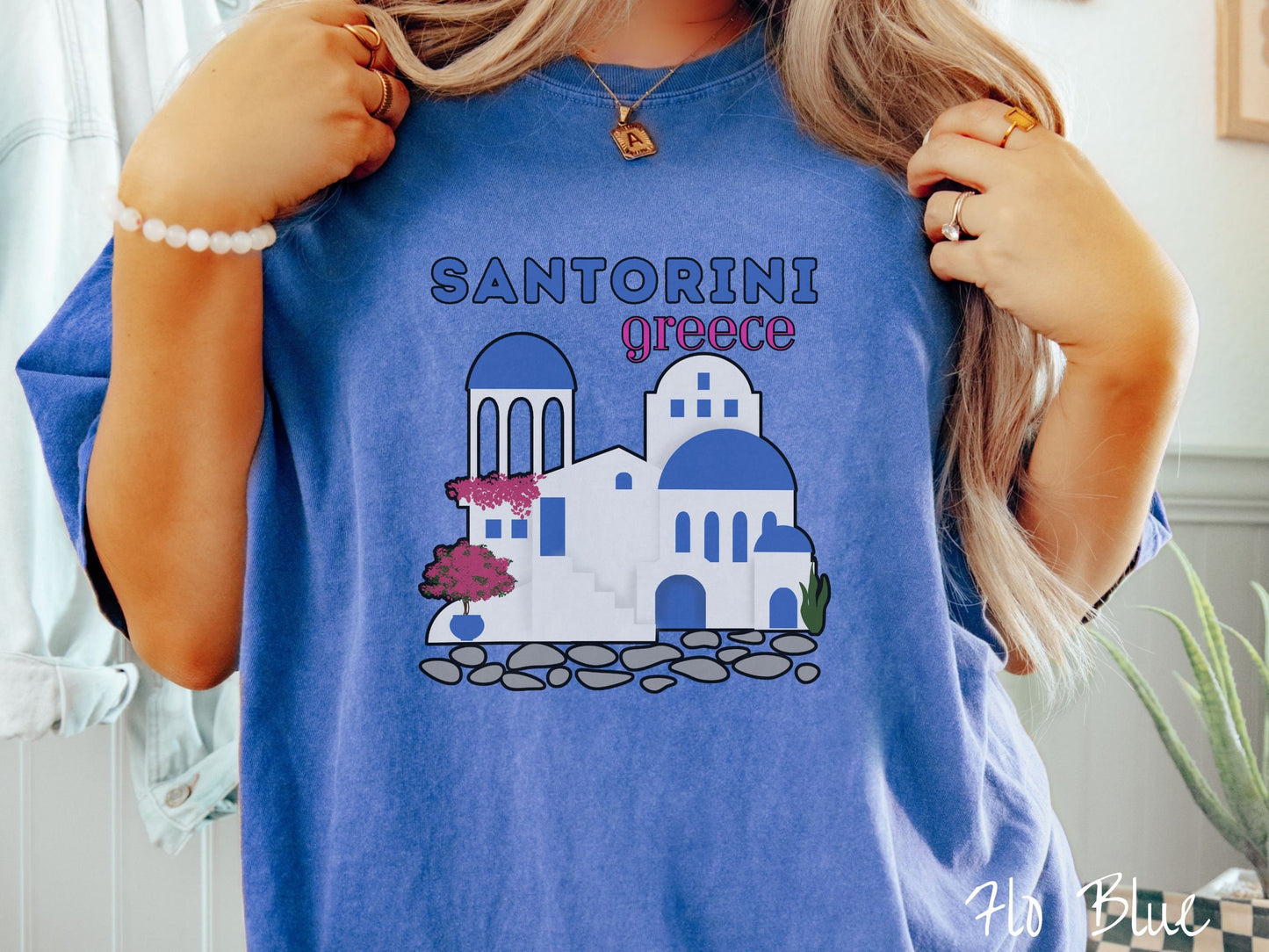 A woman wearing a cute, vintage flo blue colored Comfort Colors t-shirt with text Santorini Greece in blue and pink font. Below that is a picture of the iconic cave houses with white walls and blue trim, with cobble-stone roads and red trees.