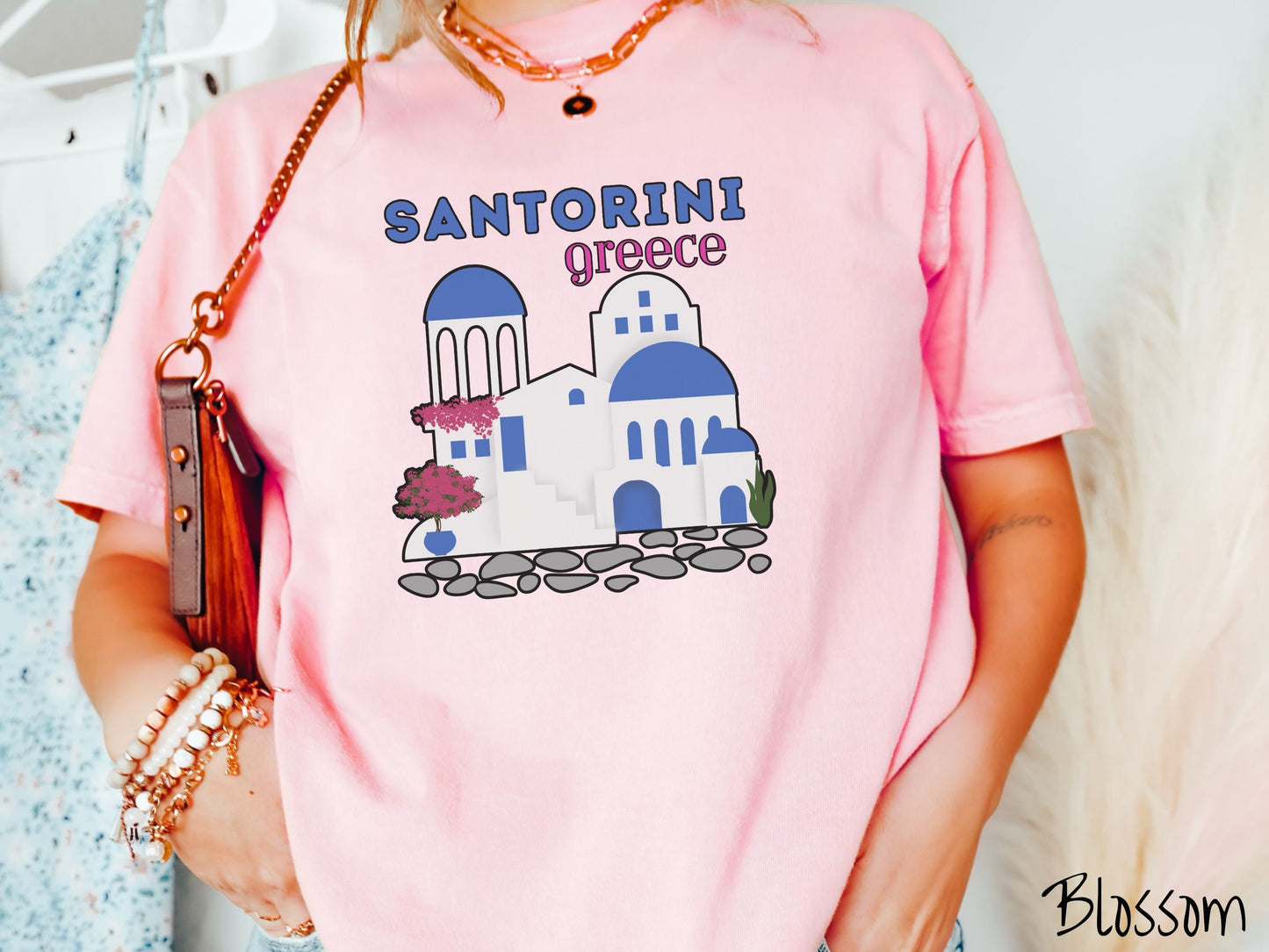 A woman wearing a cute, vintage blossom colored Comfort Colors t-shirt with text Santorini Greece in blue and pink font. Below that is a picture of the iconic cave houses with white walls and blue trim, with cobble-stone roads and red trees.