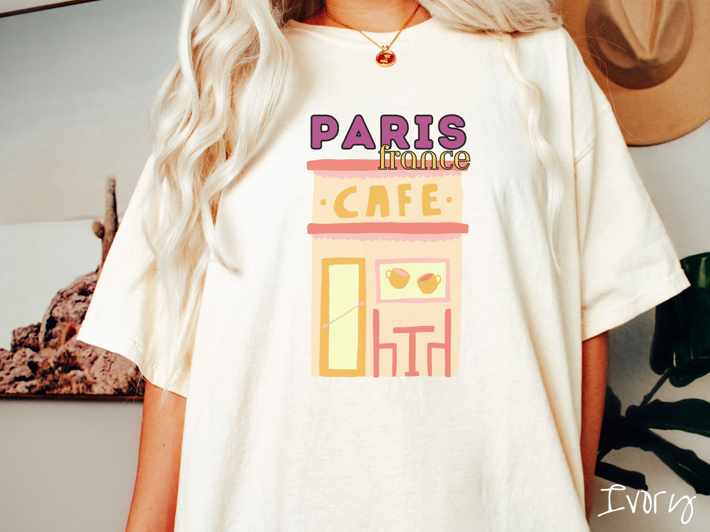 A woman wearing a cute, vintage ivory colored Comfort Colors t-shirt with the text Paris France in purpple and yellow font. Below this is a French cafe with a coffee cup picture on the outside above a table and chairs for outside seating.