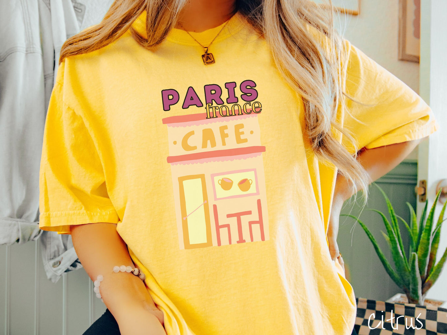 A woman wearing a cute, vintage citrus colored Comfort Colors t-shirt with the text Paris France in purpple and yellow font. Below this is a French cafe with a coffee cup picture on the outside above a table and chairs for outside seating.