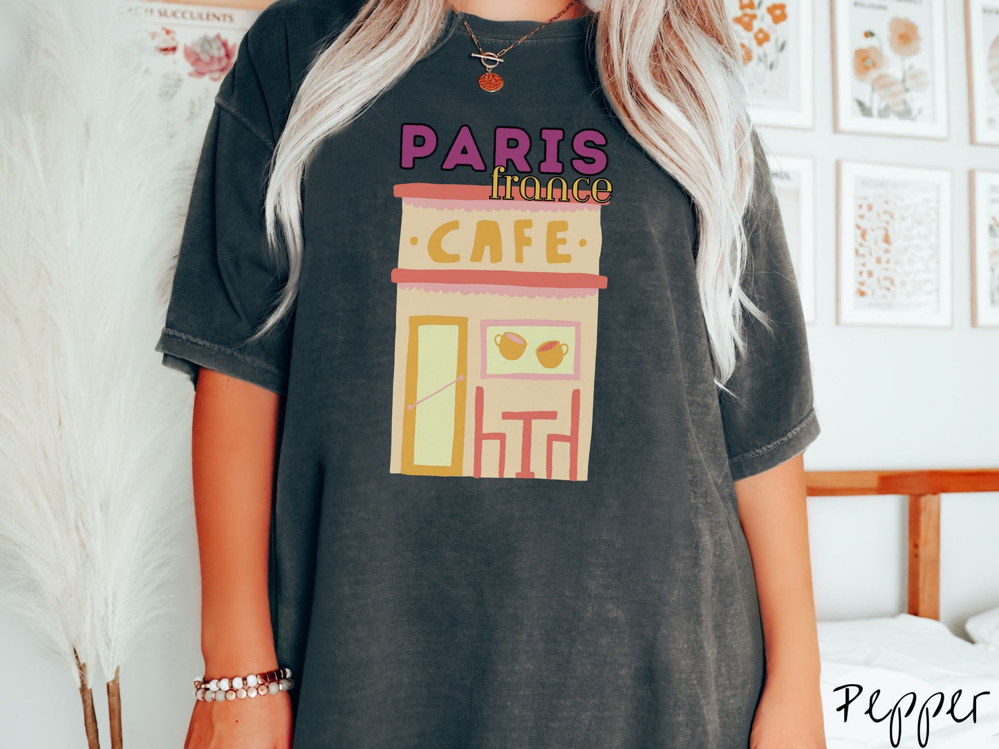 A woman wearing a cute, vintage pepper colored Comfort Colors t-shirt with the text Paris France in purpple and yellow font. Below this is a French cafe with a coffee cup picture on the outside above a table and chairs for outside seating.