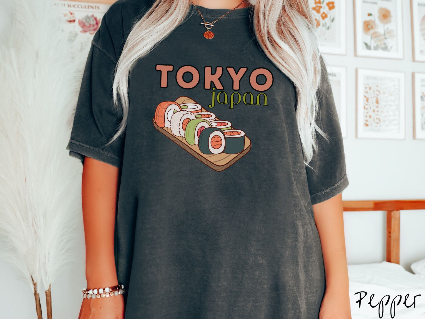 A woman wearing a vintage, pepper colored shirt with the text Tokyo Japan across the top in salmon and green font, respectively, and below is an assortment of colorful sushi rolls on a narrow wooden plate.