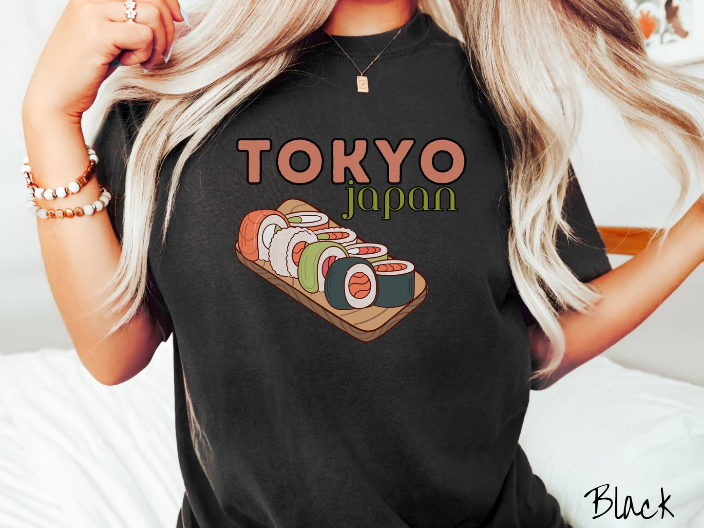 A woman wearing a vintage, black colored shirt with the text Tokyo Japan across the top in salmon and green font, respectively, and below is an assortment of colorful sushi rolls on a narrow wooden plate.