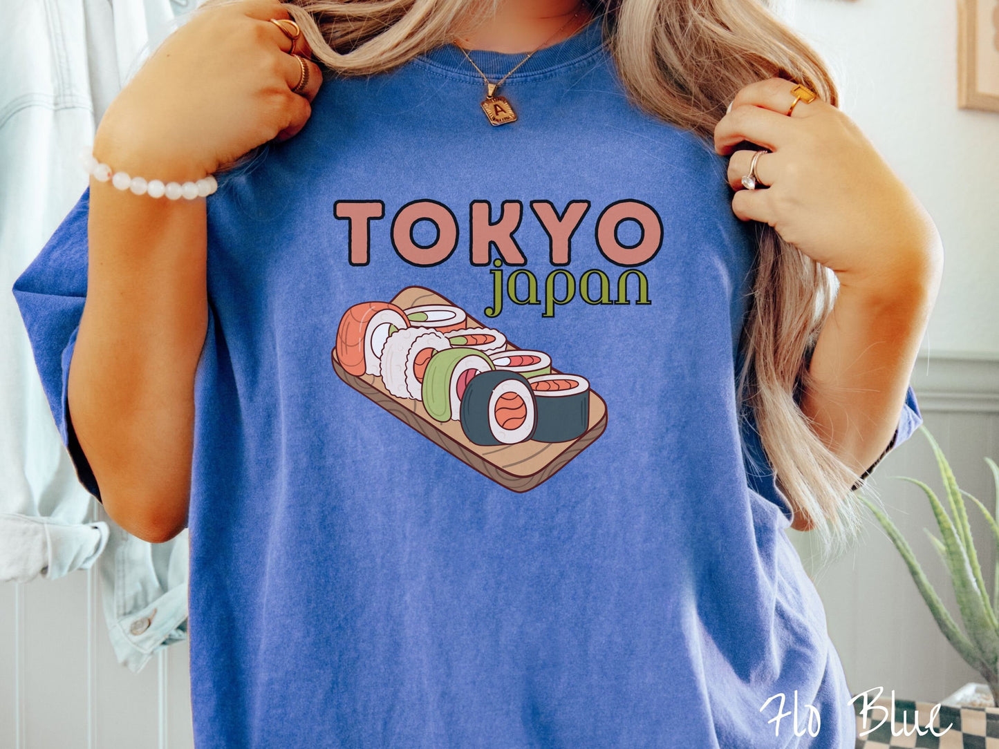 A woman wearing a vintage, flo blue colored shirt with the text Tokyo Japan across the top in salmon and green font, respectively, and below is an assortment of colorful sushi rolls on a narrow wooden plate.
