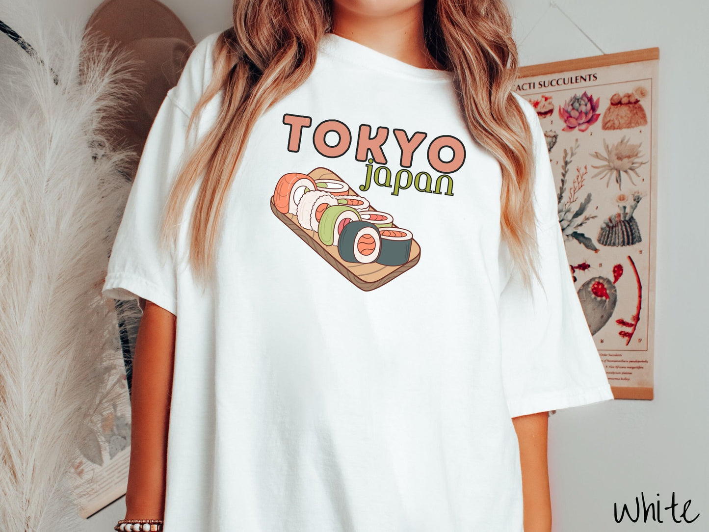 A woman wearing a vintage, white colored shirt with the text Tokyo Japan across the top in salmon and green font, respectively, and below is an assortment of colorful sushi rolls on a narrow wooden plate.