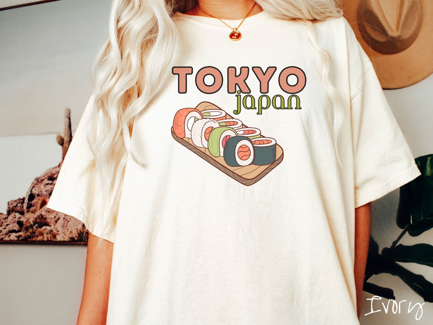 A woman wearing a vintage, ivory colored shirt with the text Tokyo Japan across the top in salmon and green font, respectively, and below is an assortment of colorful sushi rolls on a narrow wooden plate.