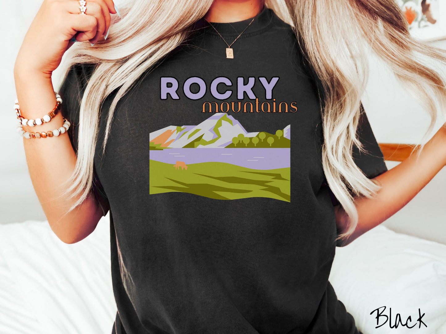 A woman wearing a vintage, black colored shirt with the text Rocky Mountains at the top and below is a scene of a massive river running beneath snowy mountains in the distance, a lone brown cow can be seen grazing in the grass next to the water.