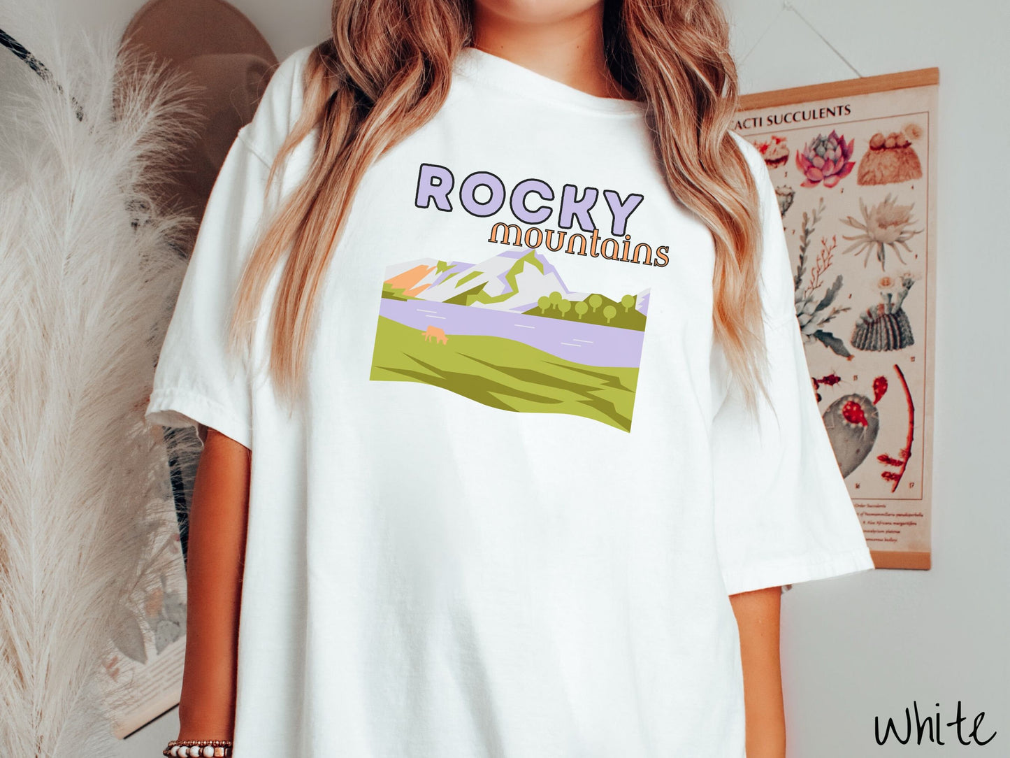 A woman wearing a vintage, white colored shirt with the text Rocky Mountains at the top and below is a scene of a massive river running beneath snowy mountains in the distance, a lone brown cow can be seen grazing in the grass next to the water.