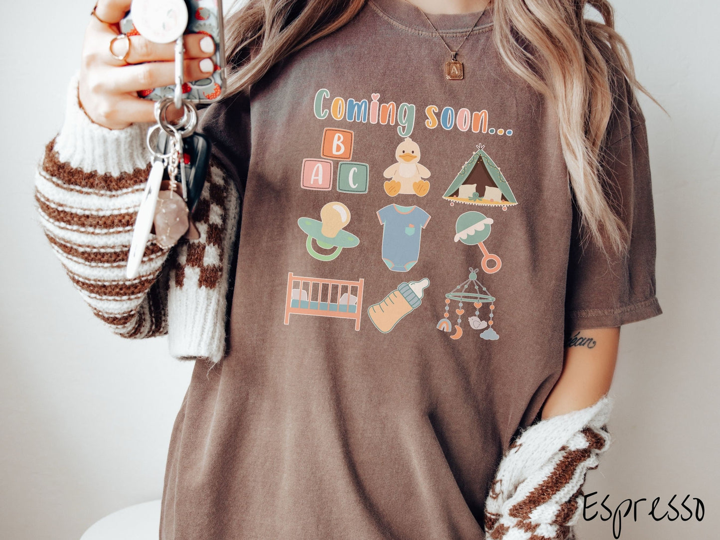 A woman wearing a vintage, espresso colored Comfort Colors t-shirt with the text in colorful font Coming soon… Below this are pictures in a three by three grid of kids toys, binkies, onesies, bottles, a crib, and alphabet blocks.