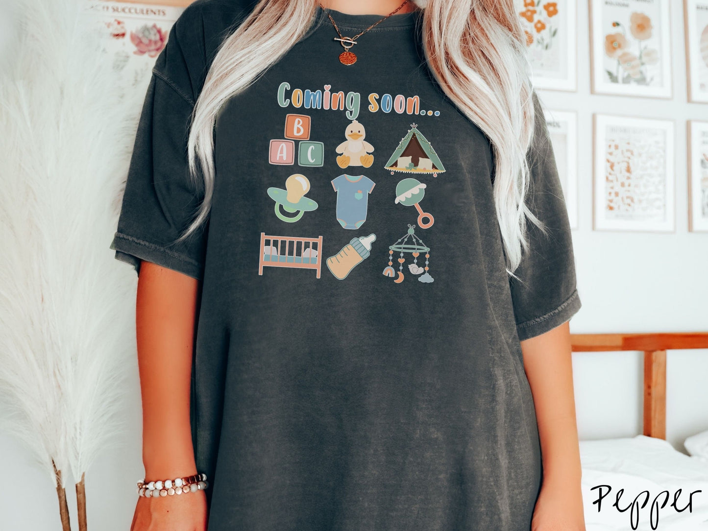 A woman wearing a vintage, pepper colored Comfort Colors t-shirt with the text in colorful font Coming soon… Below this are pictures in a three by three grid of kids toys, binkies, onesies, bottles, a crib, and alphabet blocks.