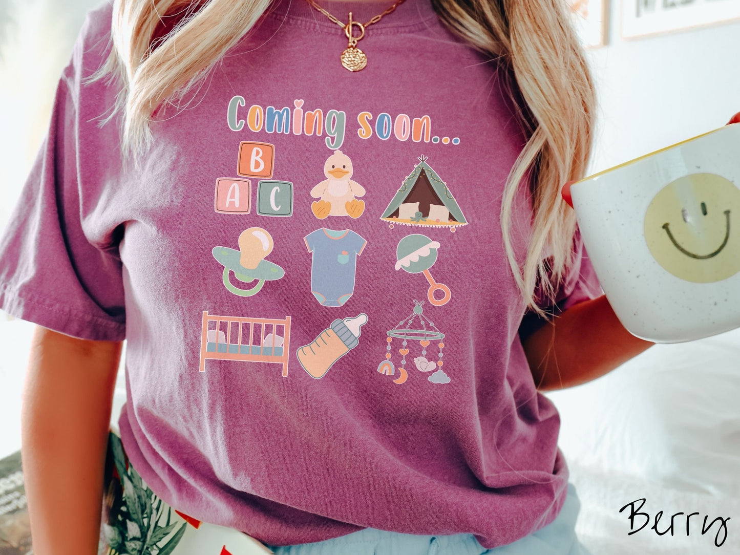 A woman wearing a vintage, berry colored Comfort Colors t-shirt with the text in colorful font Coming soon… Below this are pictures in a three by three grid of kids toys, binkies, onesies, bottles, a crib, and alphabet blocks.