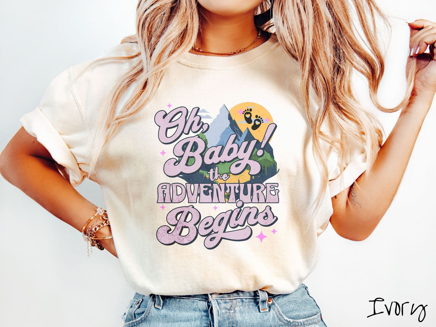 A woman wearing a vintage, ivory colored Comfort Colors t-shirt with the text in purple font Oh, Baby! The Adventure Begins! Behind this text are a blue mountain above green meadows, and behind is a yellow moon with black baby footprints inside.
