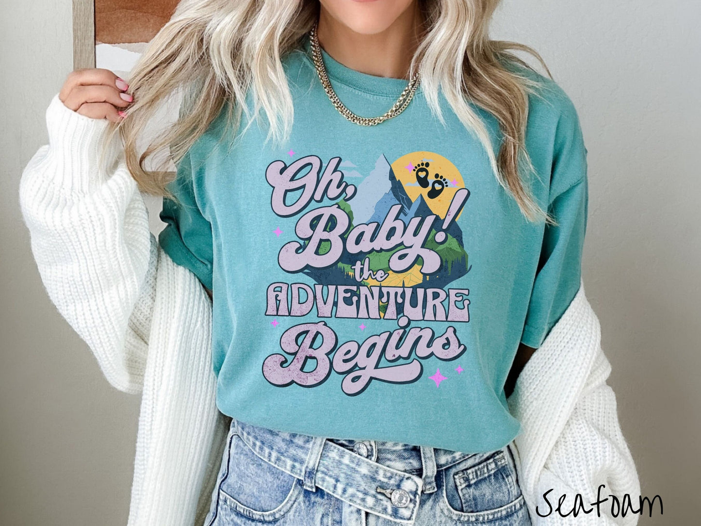A woman wearing a vintage, seafoam colored Comfort Colors t-shirt with the text in purple font Oh, Baby! The Adventure Begins! Behind this text are a blue mountain above green meadows, and behind is a yellow moon with black baby footprints inside.
