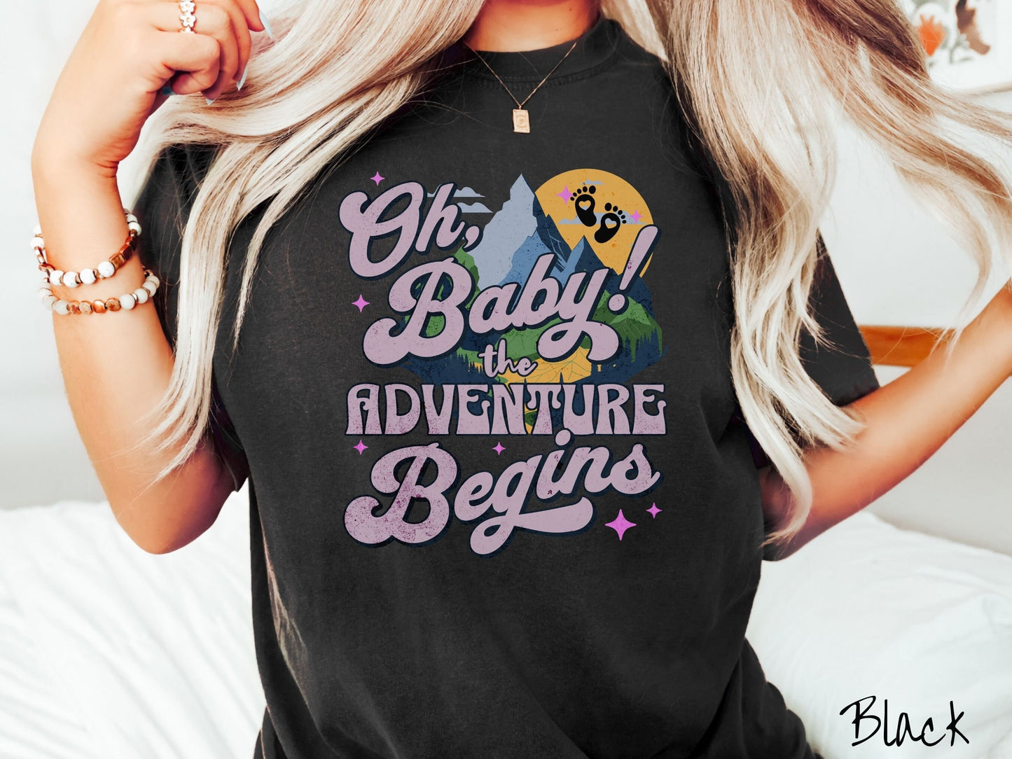 A woman wearing a vintage, black colored Comfort Colors t-shirt with the text in purple font Oh, Baby! The Adventure Begins! Behind this text are a blue mountain above green meadows, and behind is a yellow moon with black baby footprints inside.