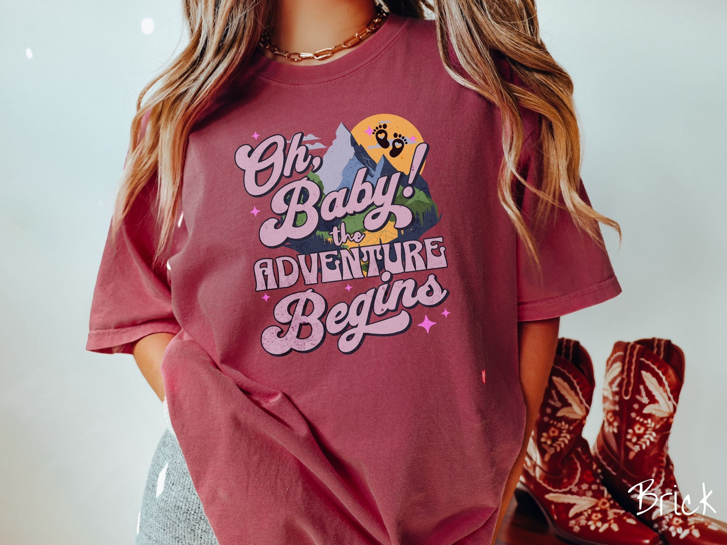 A woman wearing a vintage, brick colored Comfort Colors t-shirt with the text in purple font Oh, Baby! The Adventure Begins! Behind this text are a blue mountain above green meadows, and behind is a yellow moon with black baby footprints inside.