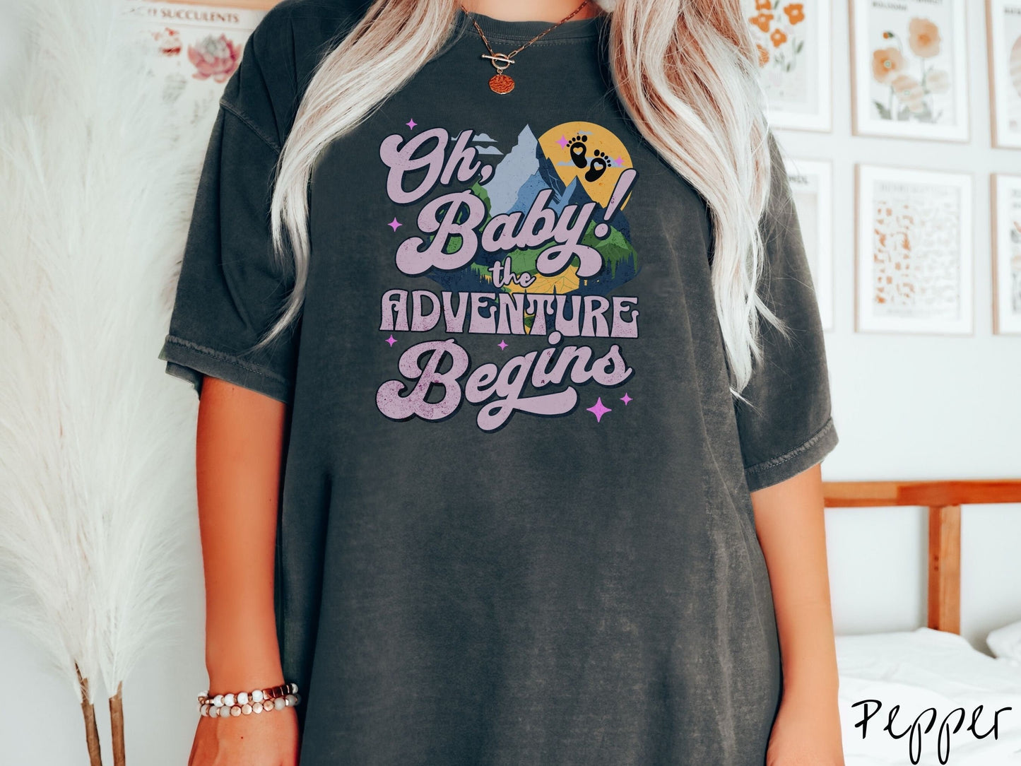 A woman wearing a vintage, pepper colored Comfort Colors t-shirt with the text in purple font Oh, Baby! The Adventure Begins! Behind this text are a blue mountain above green meadows, and behind is a yellow moon with black baby footprints inside.