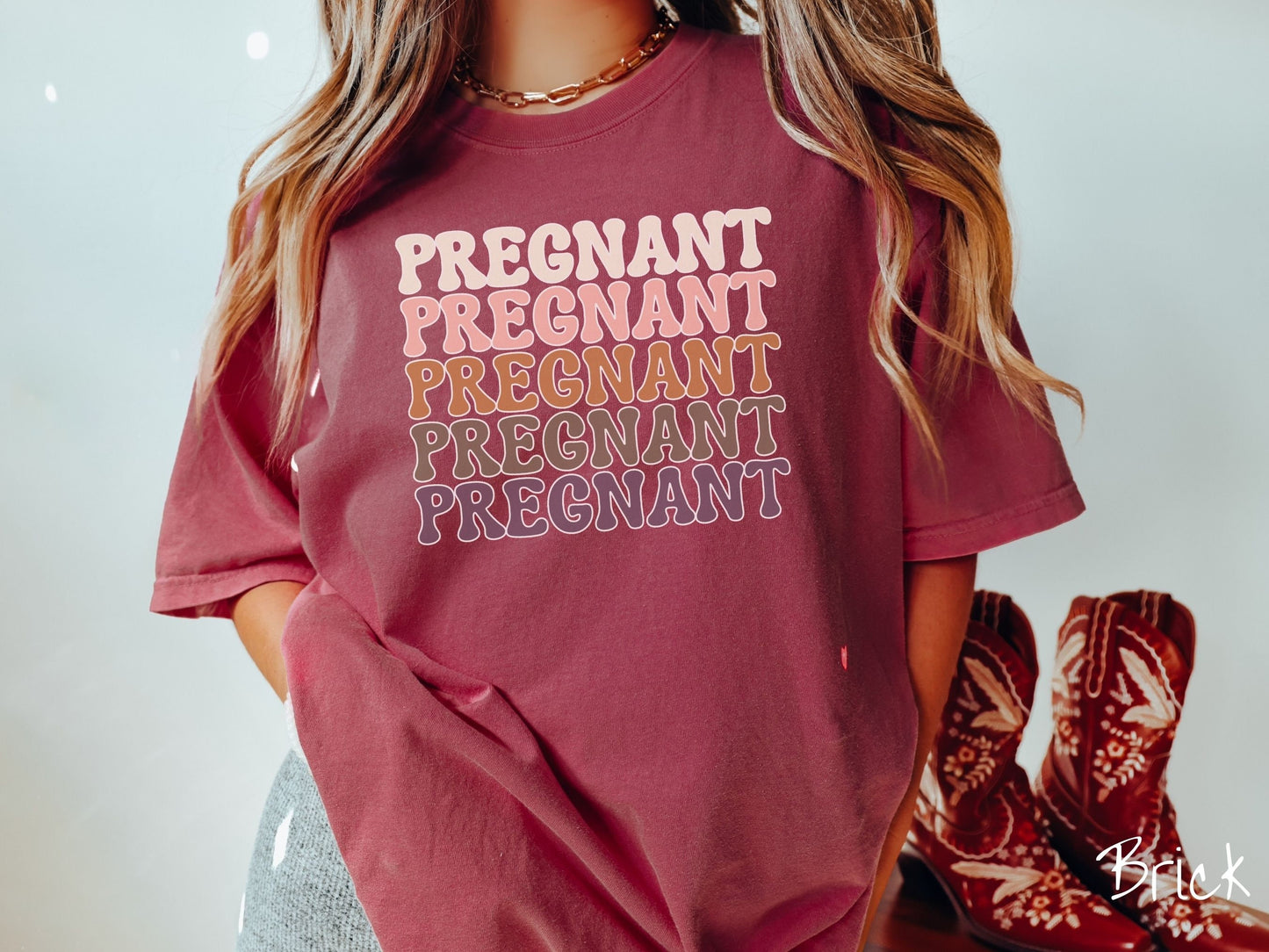 A woman wearing a vintage, brick colored Comfort Colors t-shirt with the text PREGNANT in uppercase font listed five times vertically. One is ivory colored, the next pink, next light brown, next dark brown, and the fifth is purple.