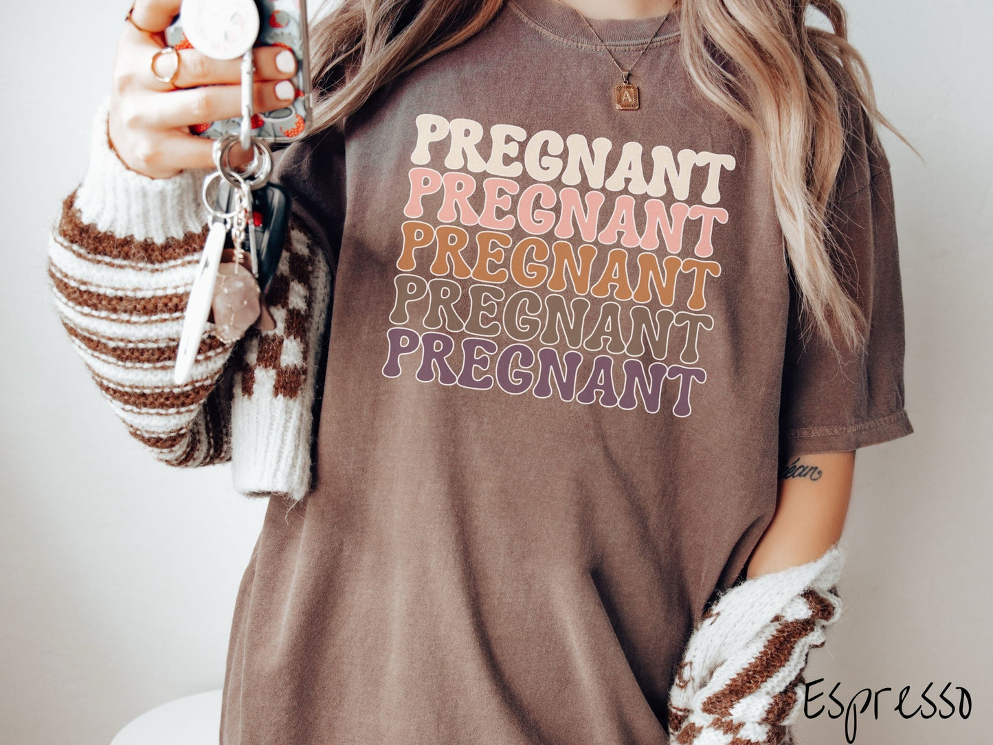 A woman wearing a vintage, espresso colored Comfort Colors t-shirt with the text PREGNANT in uppercase font listed five times vertically. One is ivory colored, the next pink, next light brown, next dark brown, and the fifth is purple.