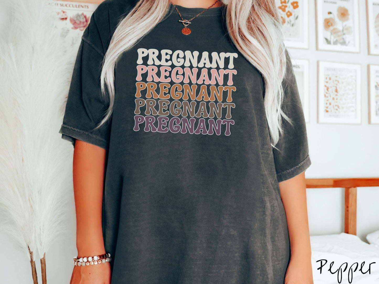 A woman wearing a vintage, pepper colored Comfort Colors t-shirt with the text PREGNANT in uppercase font listed five times vertically. One is ivory colored, the next pink, next light brown, next dark brown, and the fifth is purple.
