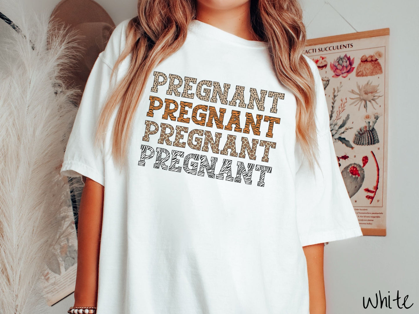 A woman wearing a vintage, white colored Comfort Colors t-shirt with the text PREGNANT in uppercase font listed four times vertically. One has leopard font, another tiger font, another zebra stripes, and the last is zebra stripe font.