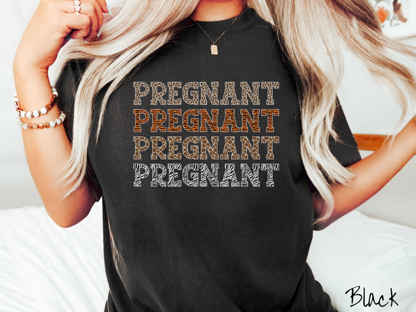 A woman wearing a vintage, black colored Comfort Colors t-shirt with the text PREGNANT in uppercase font listed four times vertically. One has leopard font, another tiger font, another zebra stripes, and the last is zebra stripe font.