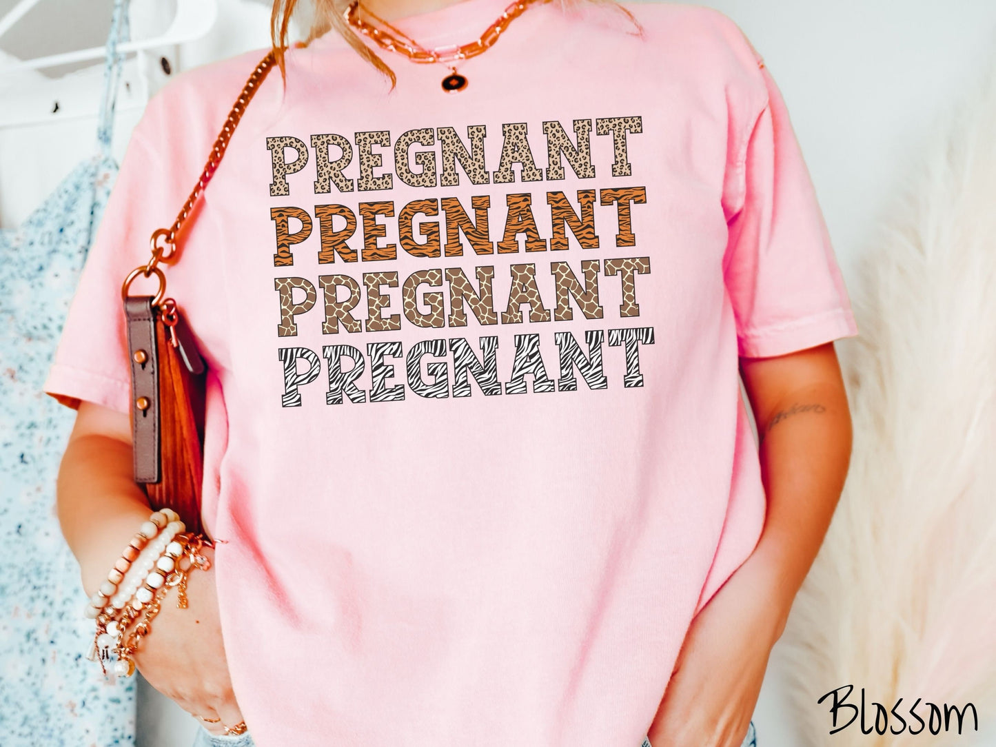 A woman wearing a vintage, blossom colored Comfort Colors t-shirt with the text PREGNANT in uppercase font listed four times vertically. One has leopard font, another tiger font, another zebra stripes, and the last is zebra stripe font.