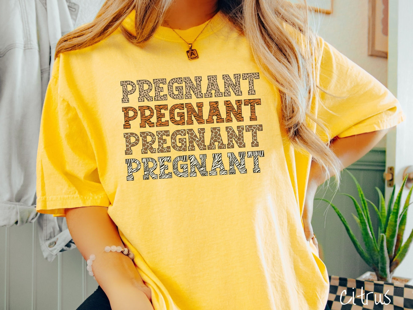 A woman wearing a vintage, citrus colored Comfort Colors t-shirt with the text PREGNANT in uppercase font listed four times vertically. One has leopard font, another tiger font, another zebra stripes, and the last is zebra stripe font.