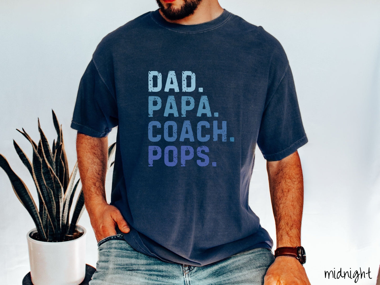 A man wearing a vintage, midnight colored Comfort Colors t-shirt with the text in varying blue colored font Dad. Papa. Coach. Pops.