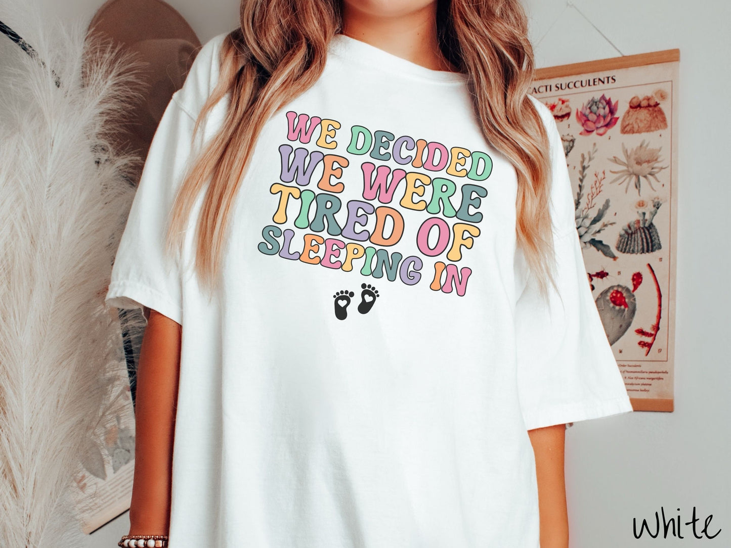 A woman wearing a vintage, white colored Comfort Colors t-shirt with the text We Decided We Were Tired Of Sleeping In in pink, yellow, green, and purple uppercase font. Below the text are two white baby footprints with hearts inside them.