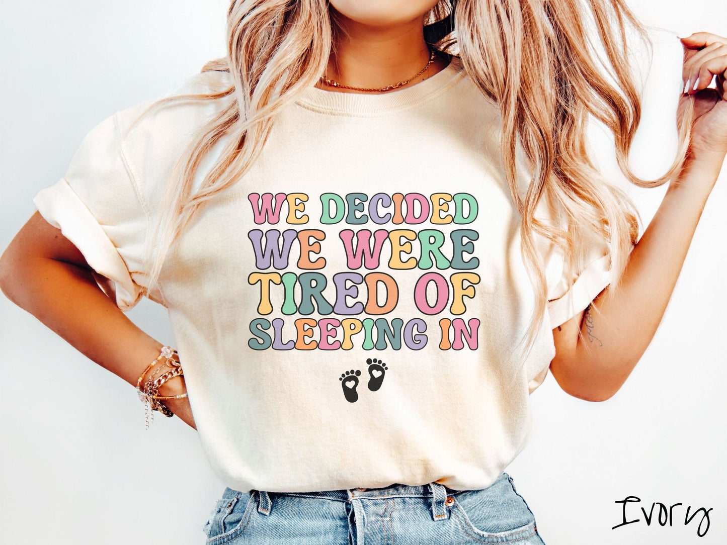 A woman wearing a vintage, ivory colored Comfort Colors t-shirt with the text We Decided We Were Tired Of Sleeping In in pink, yellow, green, and purple uppercase font. Below the text are two white baby footprints with hearts inside them.