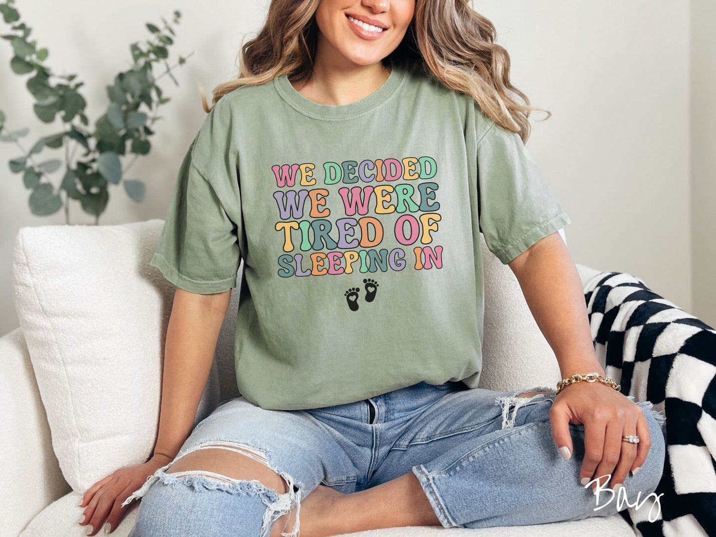 A woman wearing a vintage, bay colored Comfort Colors t-shirt with the text We Decided We Were Tired Of Sleeping In in pink, yellow, green, and purple uppercase font. Below the text are two white baby footprints with hearts inside them.