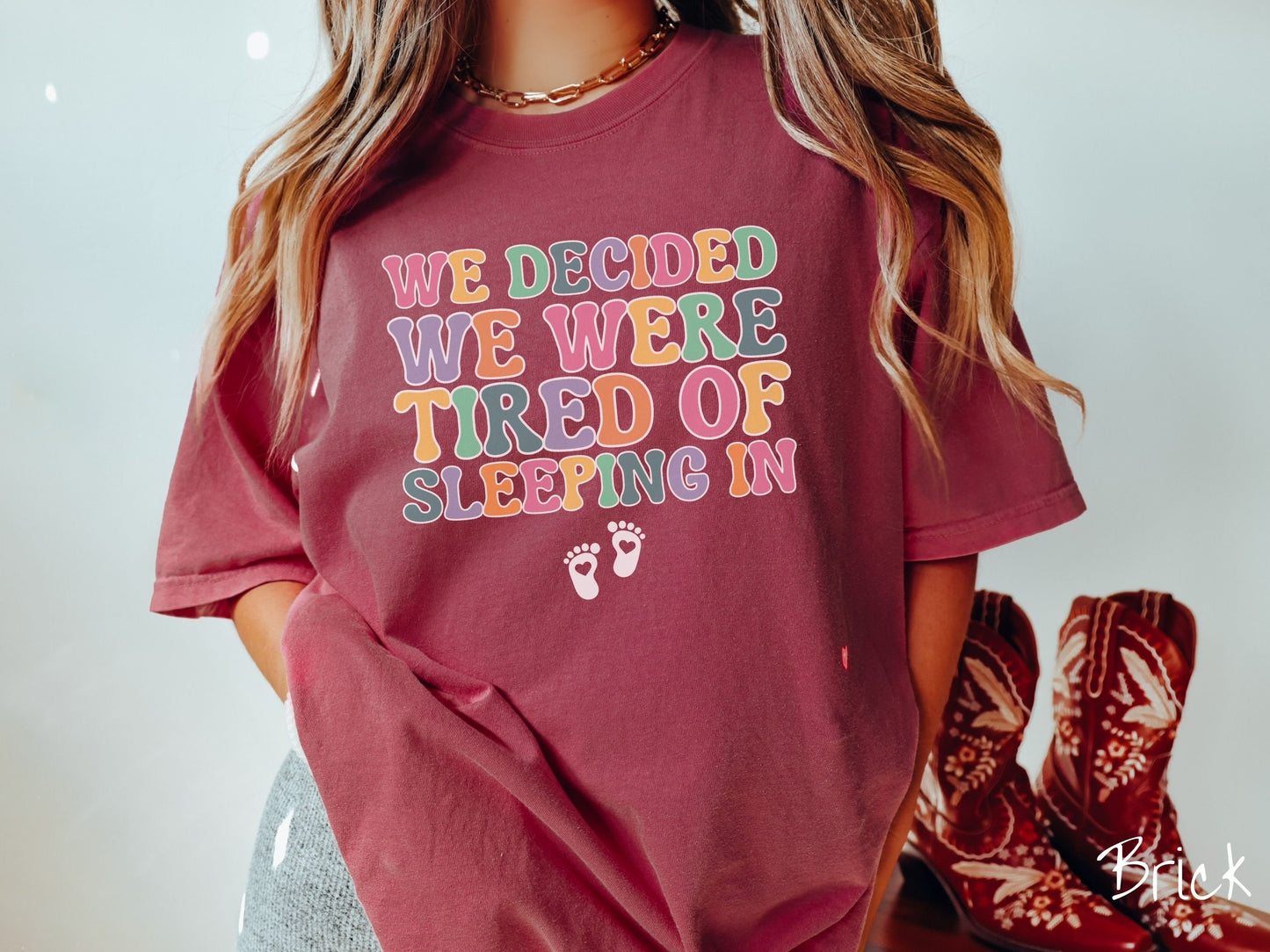 A woman wearing a vintage, brick colored Comfort Colors t-shirt with the text We Decided We Were Tired Of Sleeping In in pink, yellow, green, and purple uppercase font. Below the text are two white baby footprints with hearts inside them.