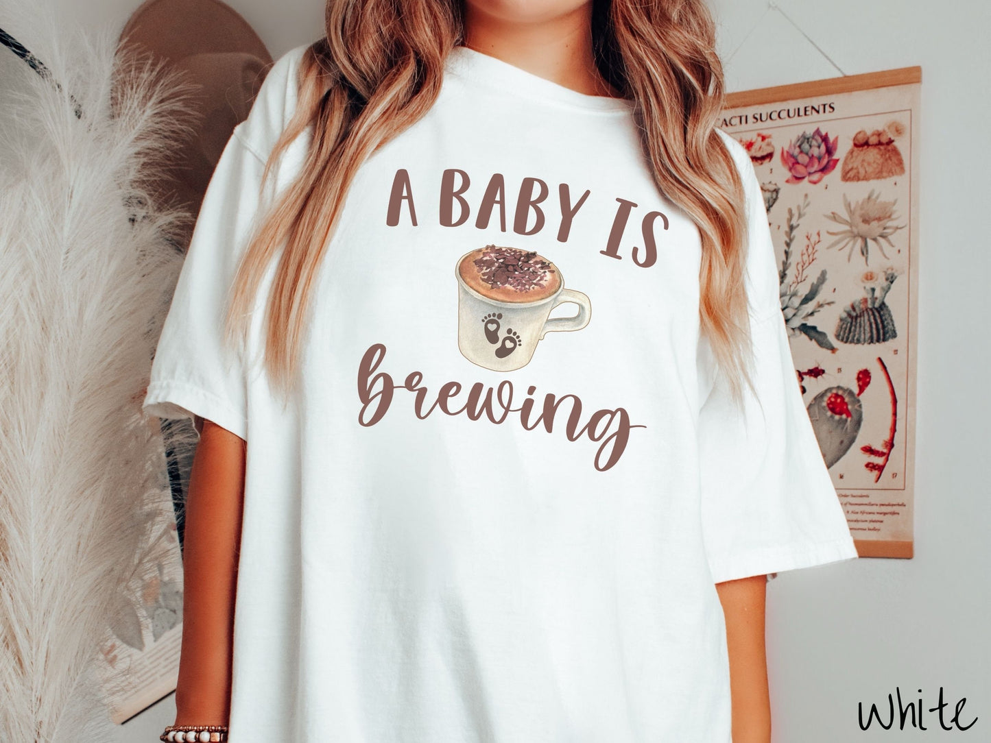 A woman wearing a vintage, white colored Comfort Colors t-shirt with the text A Baby Is Brewing in light brown font. In between the text is a white cup of coffee with cream on top. The cup has two baby footprints on it with hearts inside them.