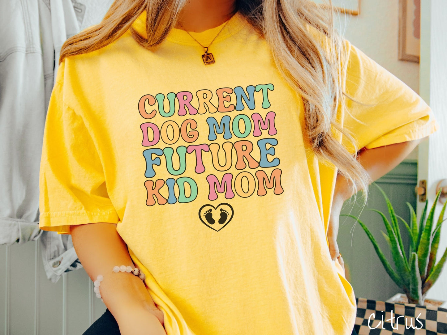 A woman wearing a vintage, citrus colored Comfort Colors t-shirt with the text Curent Dog Mom Future Kid Mom in orange, green, pink, yellow, and blue uppercase font. Below this is a black outlined heart with two kid footprints inside.