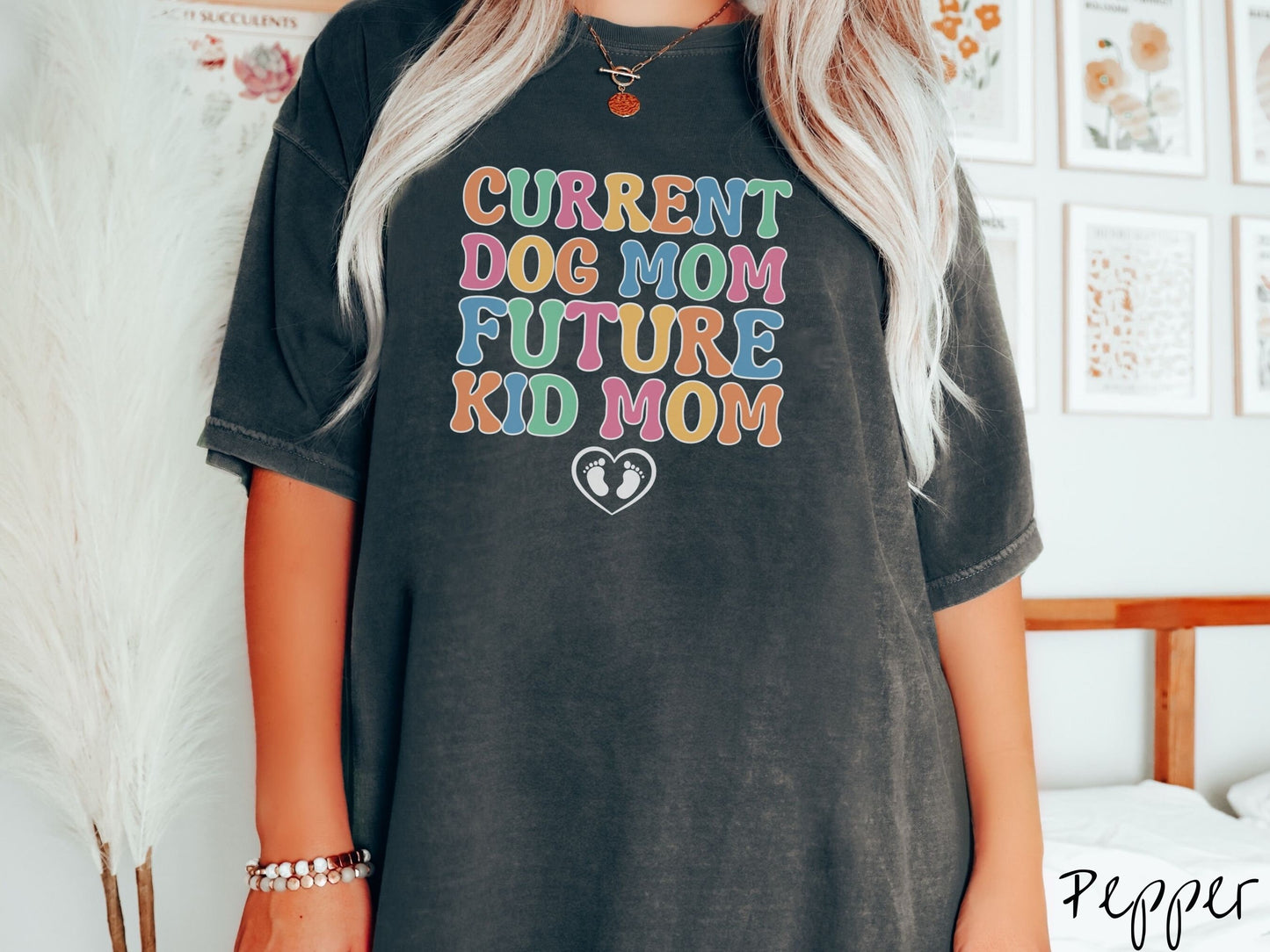 A woman wearing a vintage, pepper colored Comfort Colors t-shirt with the text Curent Dog Mom Future Kid Mom in orange, green, pink, yellow, and blue uppercase font. Below this is a black outlined heart with two kid footprints inside.