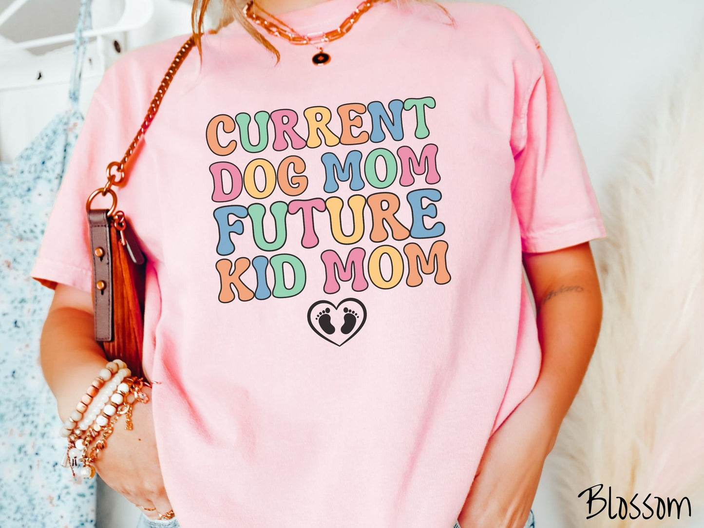 A woman wearing a vintage, blossom colored Comfort Colors t-shirt with the text Curent Dog Mom Future Kid Mom in orange, green, pink, yellow, and blue uppercase font. Below this is a black outlined heart with two kid footprints inside.
