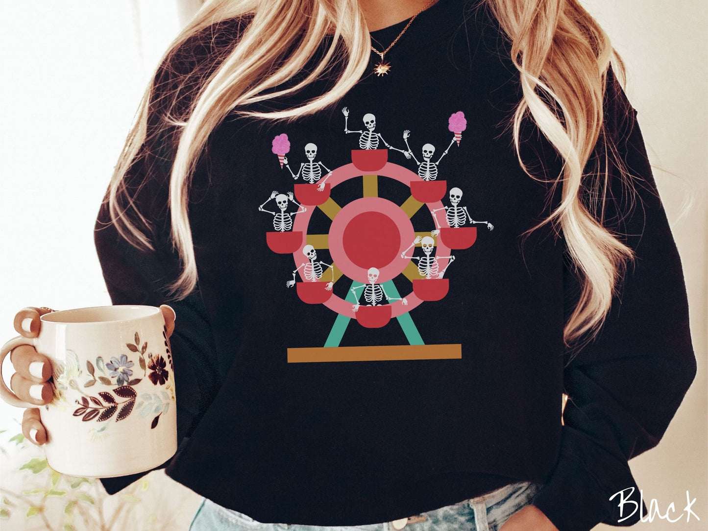 A woman wearing a cute black colored comfy sweatshirt with a ferris wheel filled with black skeletons waving and holding cotton candy as they go around the ride.