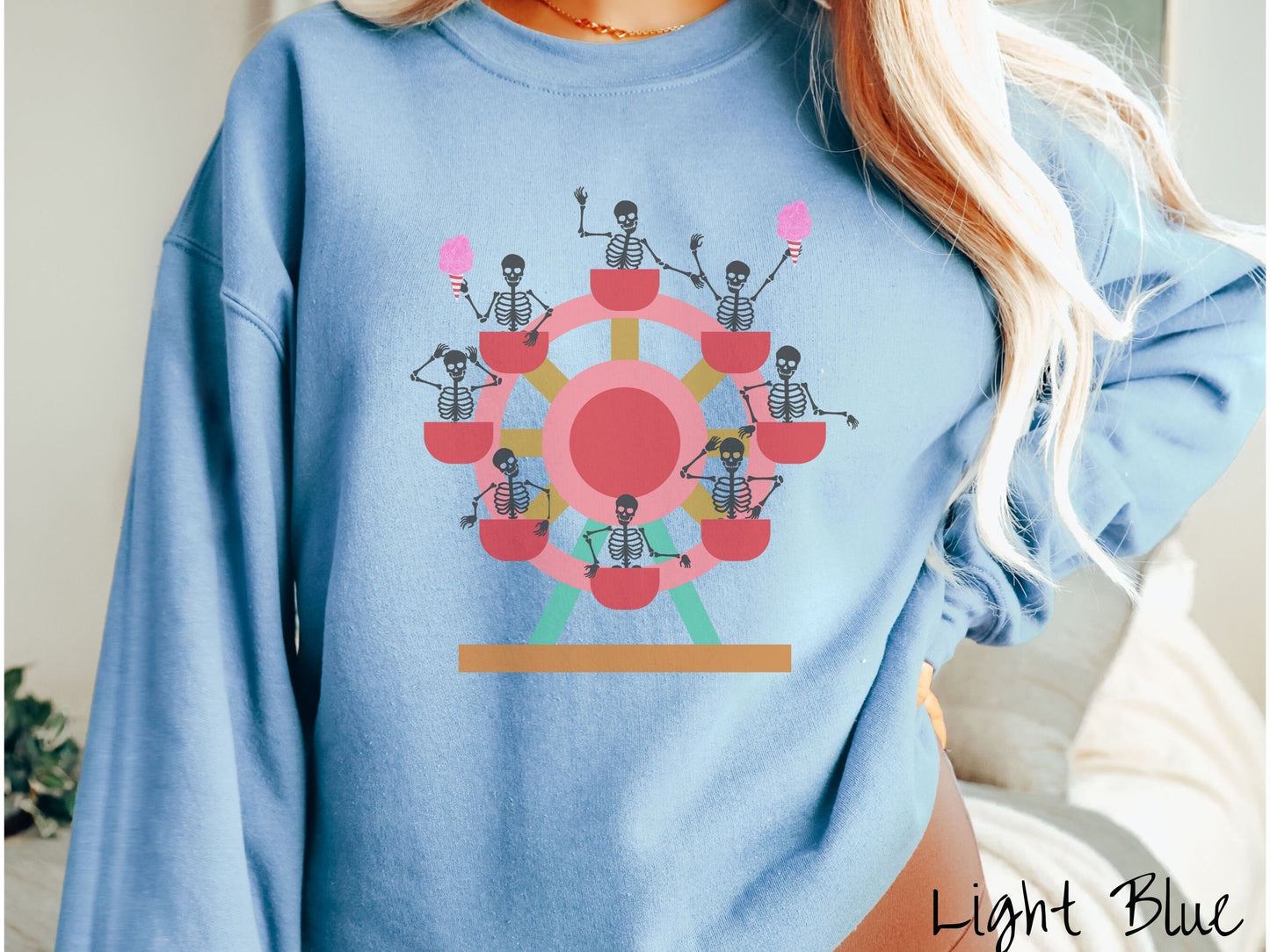 A woman wearing a cute light blue colored comfy sweatshirt with a ferris wheel filled with black skeletons waving and holding cotton candy as they go around the ride.