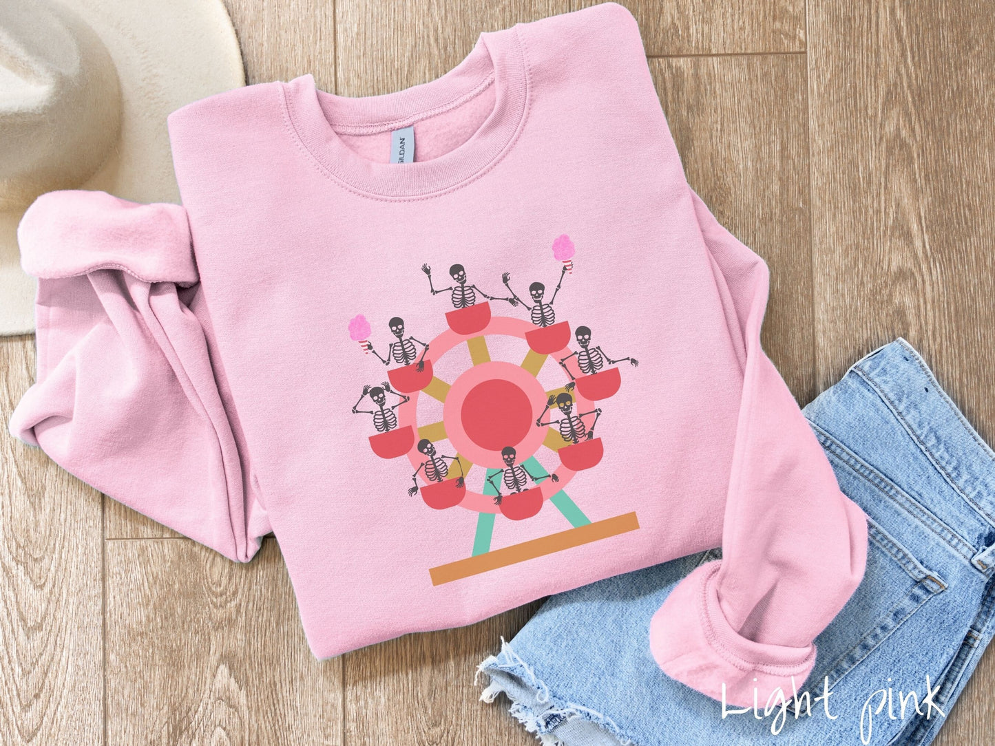 A cute light pink colored comfy sweatshirt with a ferris wheel filled with black skeletons waving and holding cotton candy as they go around the ride.