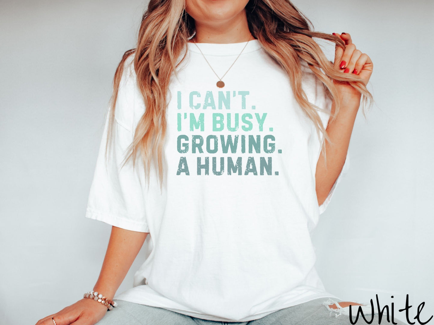 A woman wearing a vintage, white colored Comfort Colors t-shirt with the text listed vertically in varying shades of green font I Can’t. I’m busy. Growing. A human.