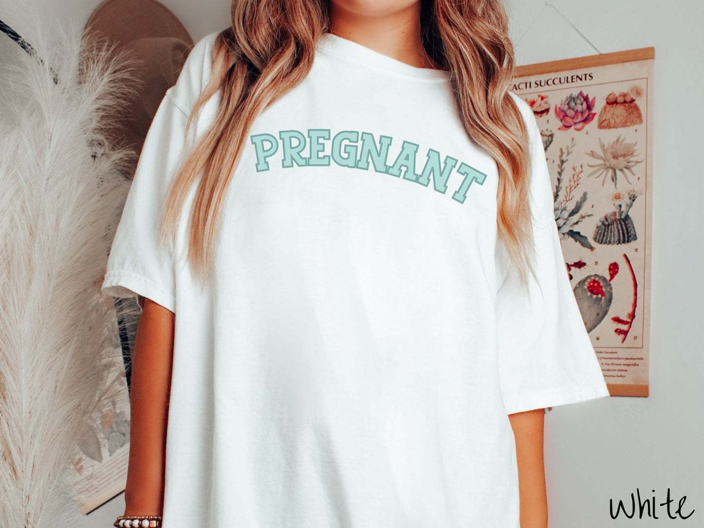 A woman wearing a vintage, white colored Comfort Colors t-shirt with the text PREGNANT in light green and dark green outlined uppercase font.