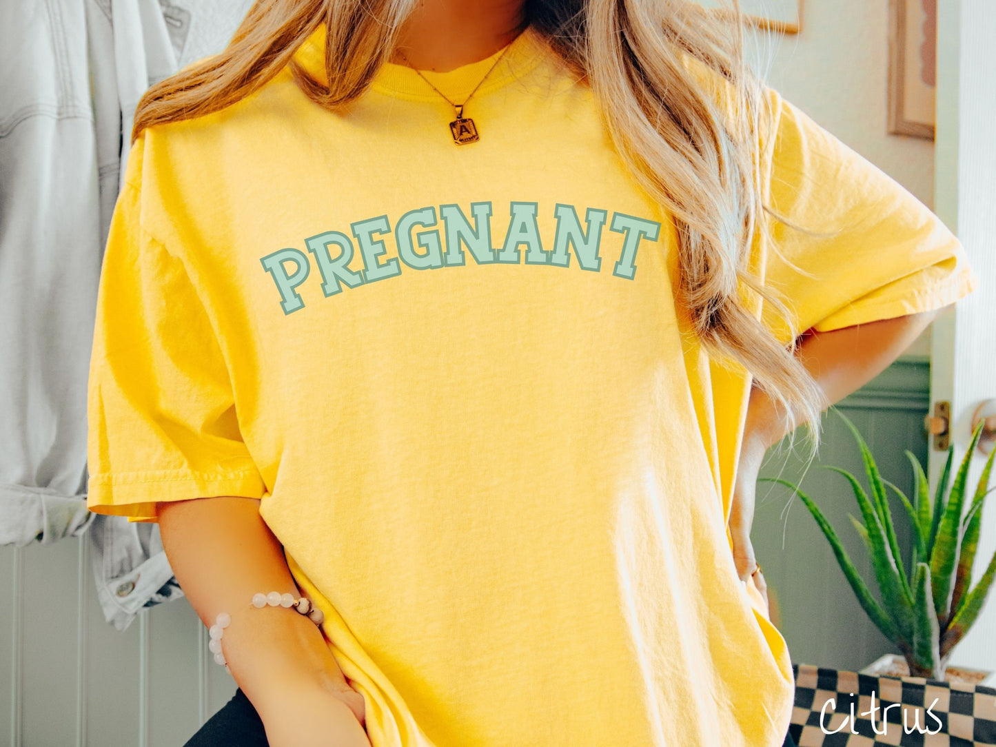 A woman wearing a vintage, citrus colored Comfort Colors t-shirt with the text PREGNANT in light green and dark green outlined uppercase font.